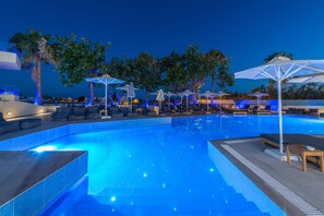 A swimming pool with lounge chairs and umbrellas, surrounded by palm trees and night lighting. - Imperial Palace Hotel (Rethymno)