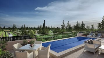 A rooftop pool area with a glass balustrade, wicker furniture, and a view of a landscaped garden.