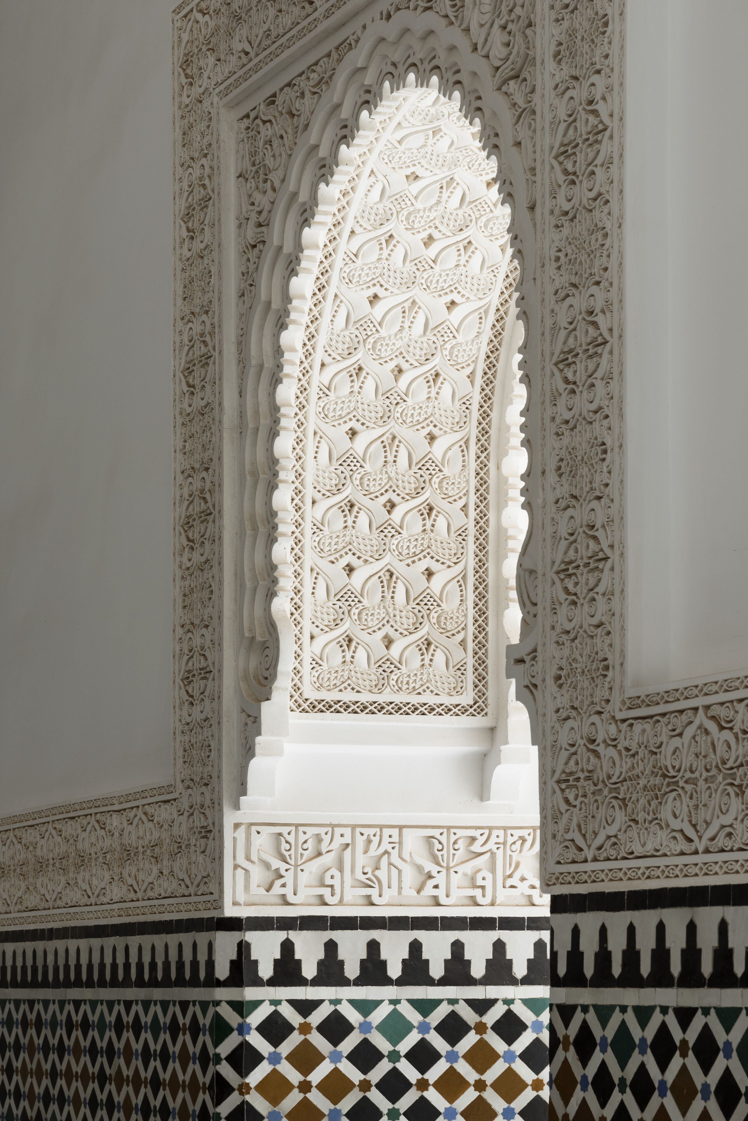 An intricately carved white archway with detailed geometric and floral patterns, set within a tiled wall with a geometric border.