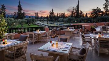 An outdoor dining area with wicker furniture and tables set for two, overlooking a landscaped garden and distant mountains.
