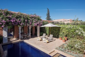 Zona de piscina con sillones de descanso, una pérgola con enredaderas floreadas y un edificio al fondo.