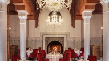 A luxurious dining area with ornate ceiling and chandelier, red chairs, and white tablecloths.