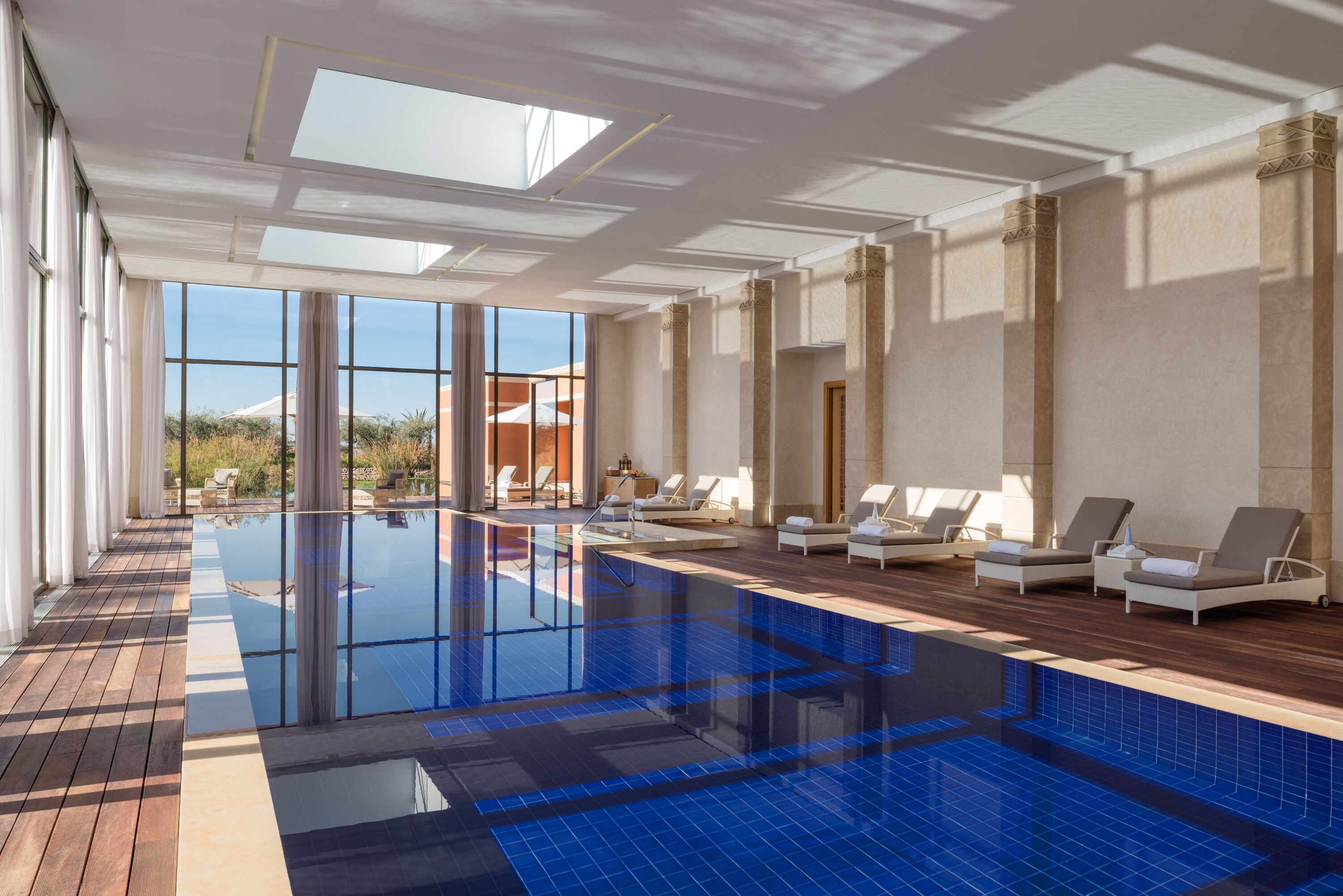 Une grande piscine intérieure avec des parois en verre transparent, une terrasse en bois et des chaises longues.