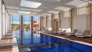 A large indoor swimming pool with clear glass walls, wooden deck, and lounge chairs.