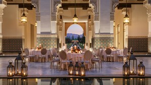 Un comedor lujoso con mesas dispuestas para una comida, columnas ornamentales y una piscina que refleja la escena.