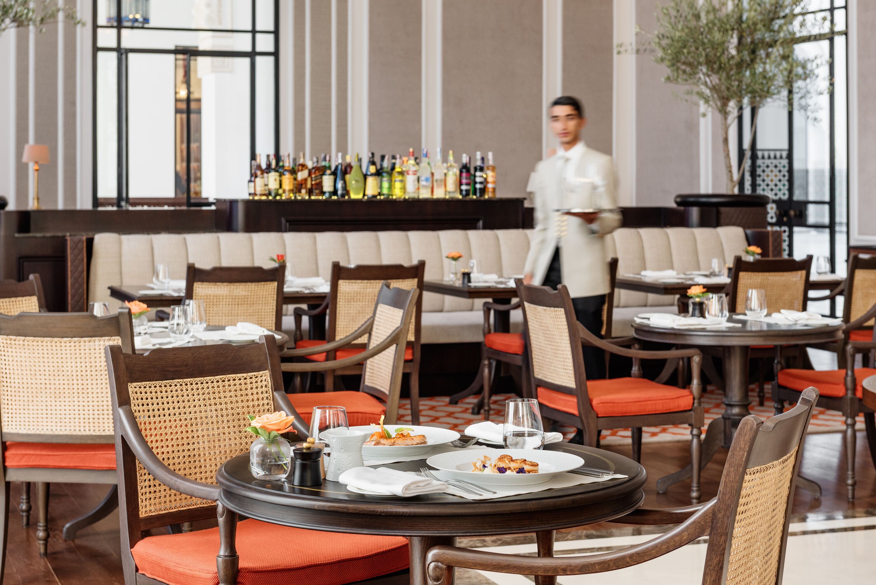 A restaurant with round tables, wicker chairs, and a bar area in the background.