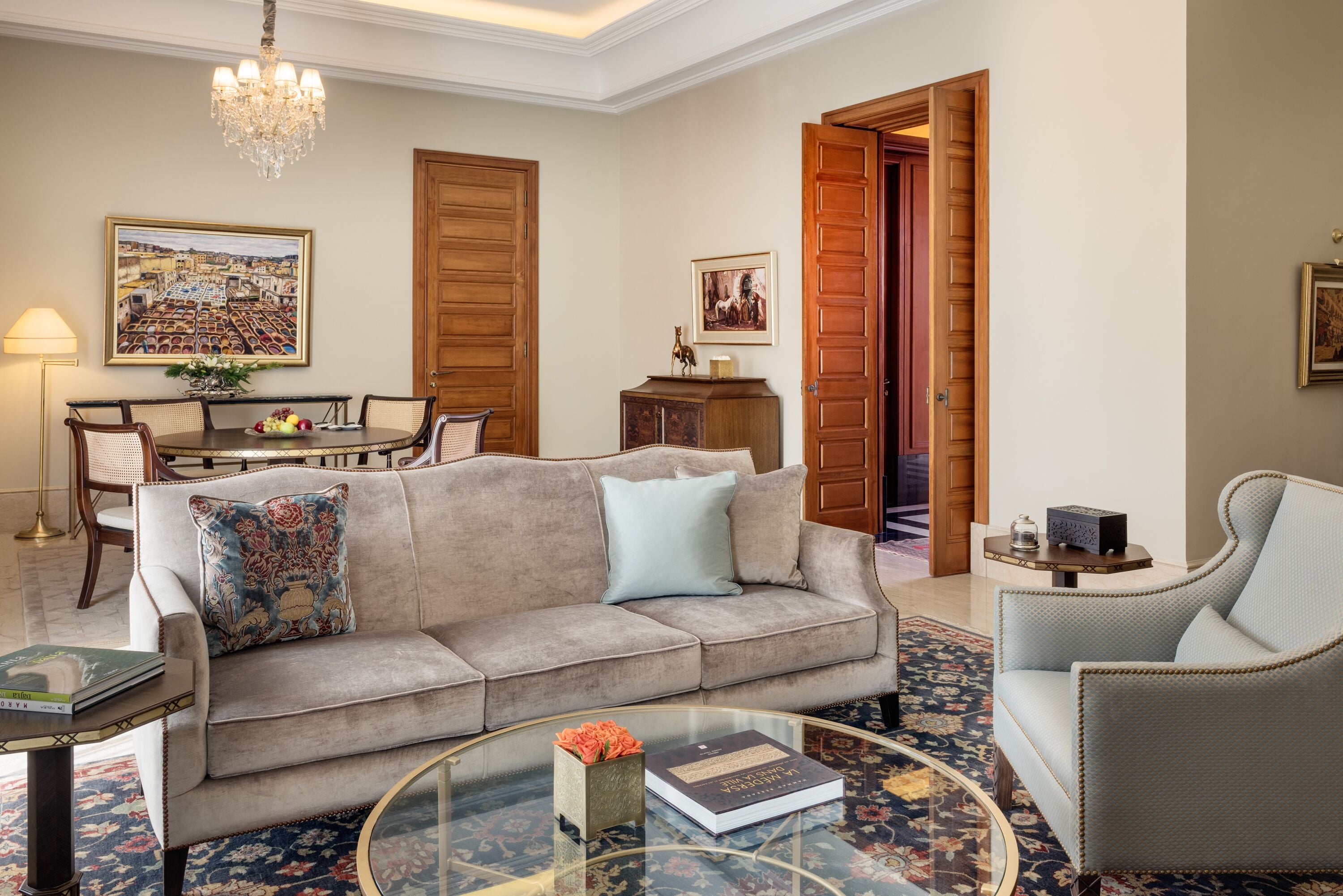A living room with a sofa, armchairs, a glass coffee table, a dining area with a table and chairs, and a chandelier.