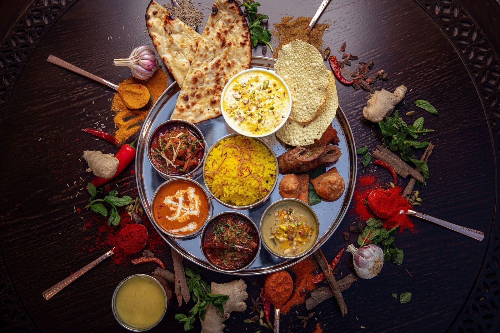 A variety of Indian dishes on a metal tray, including naan bread, curry, and rice, surrounded by spices and herbs.