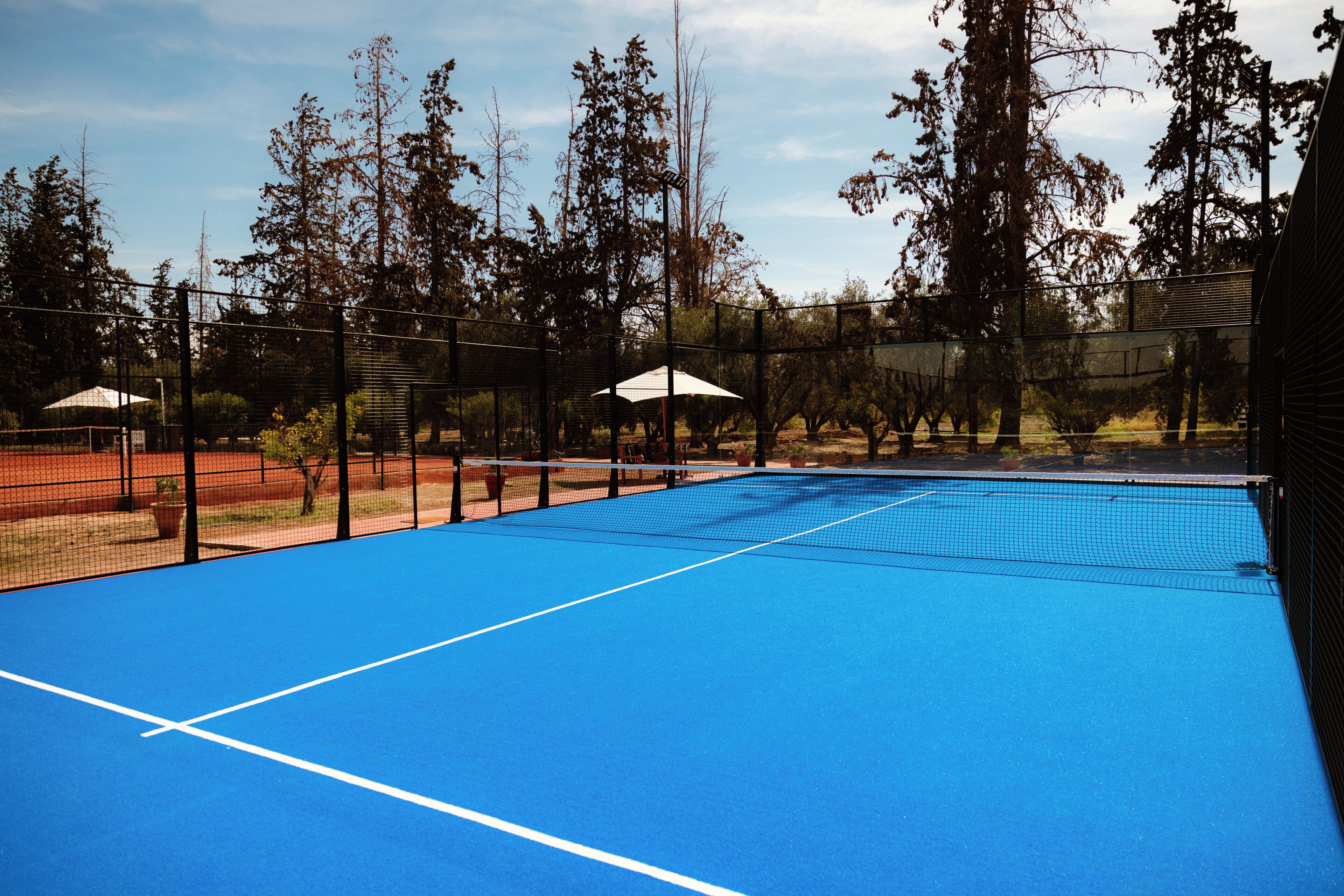 An outdoor tennis court with a blue surface, surrounded by trees and a fence.