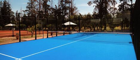 An outdoor tennis court with a blue surface, surrounded by trees and a fence.