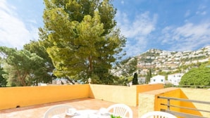 A terrace with a table set for two, surrounded by white chairs, with a view of a hillside and trees. - Velazquez by Interhome (Roses)