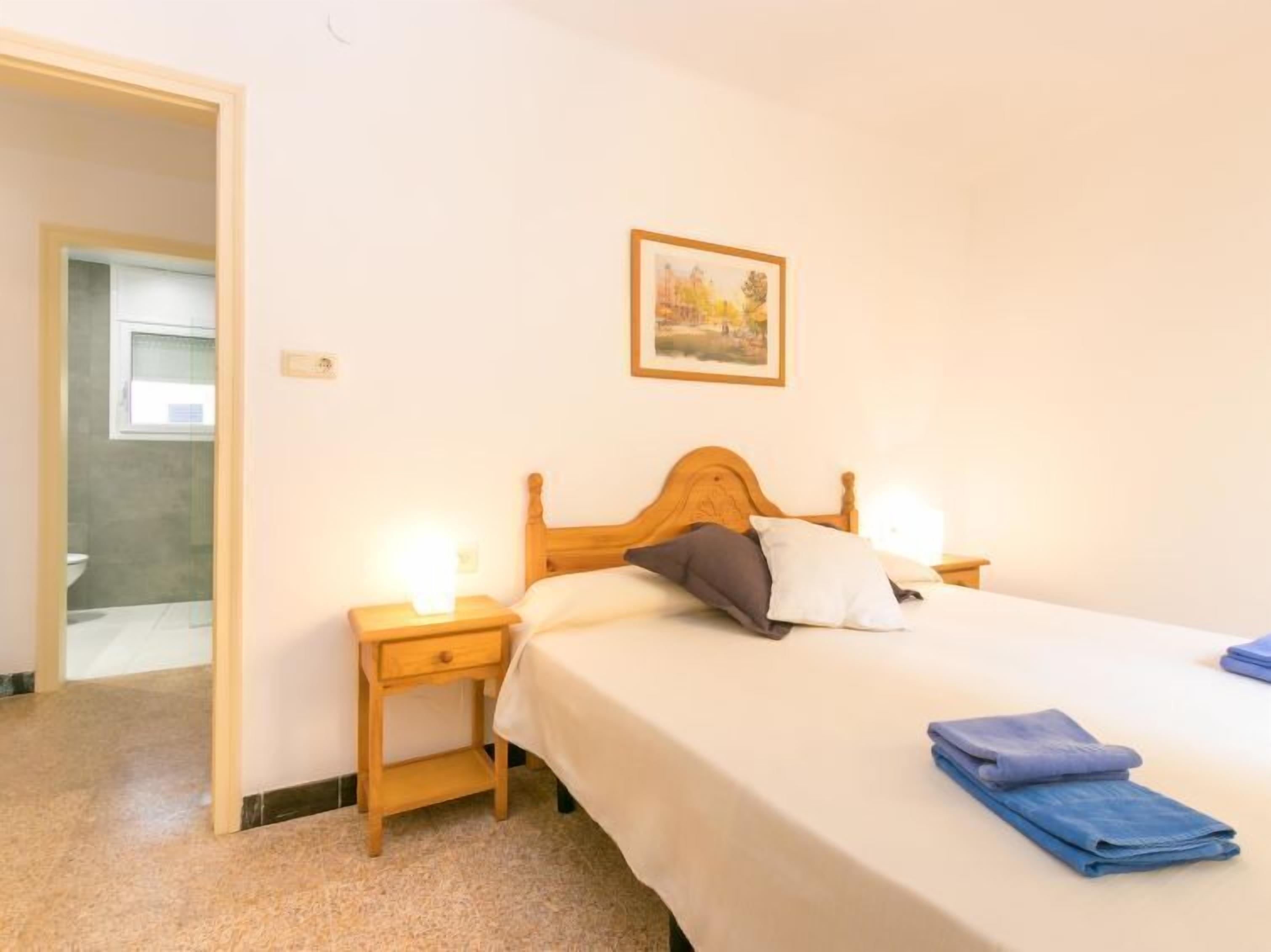 Een slaapkamer met een houten bed, twee bedlampen, een nachtkastje, een ingelijst schilderij aan de muur, en een badkamer zichtbaar door een geopende deur.