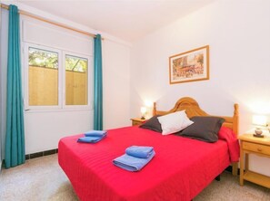 A bedroom with a red bedspread, two pillows, a wooden headboard, a nightstand with a lamp, and a window with curtains. - Velazquez by Interhome (Roses)