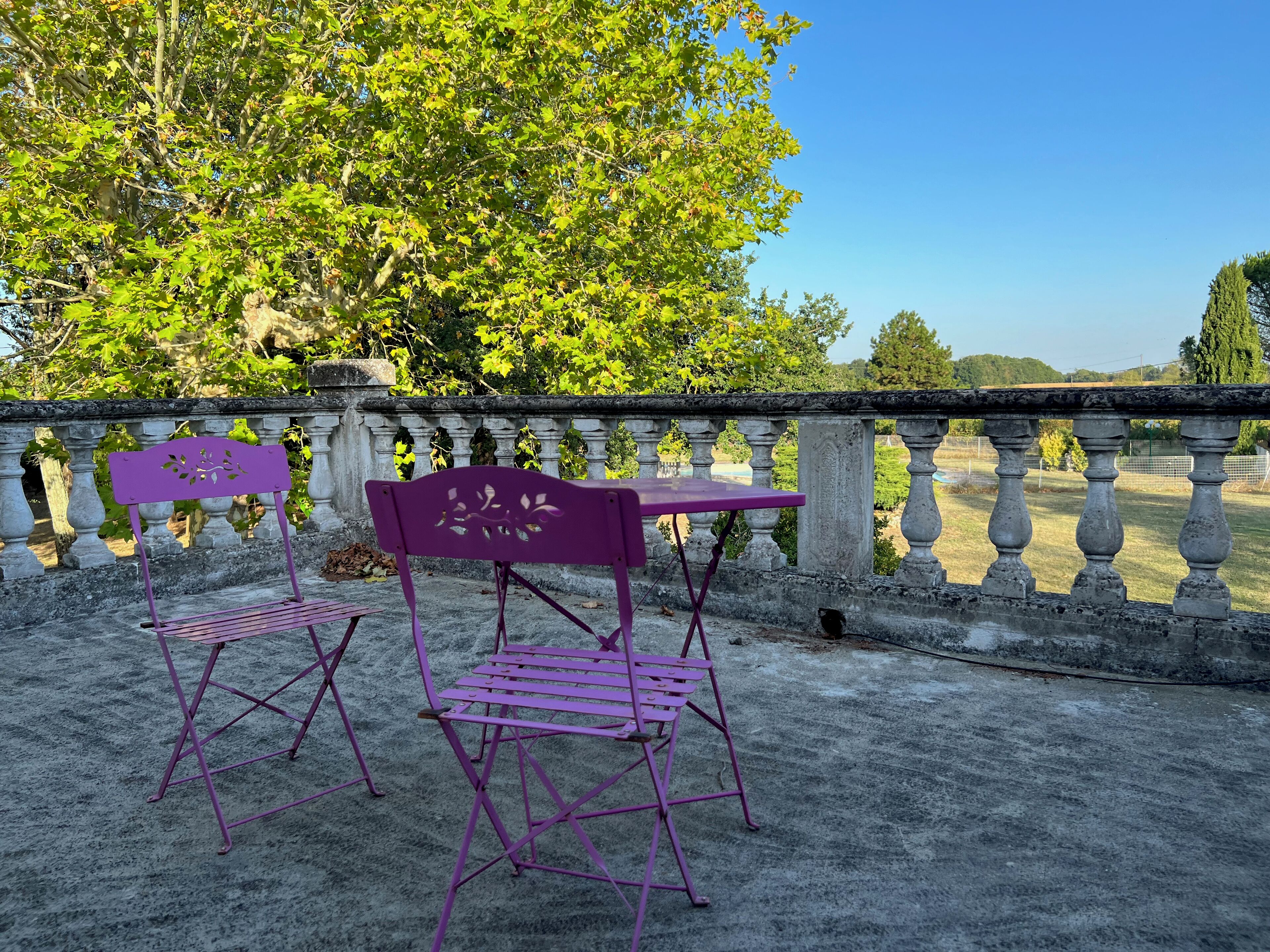 Un balcón con dos sillas moradas y una mesa, con vista a un jardín con barandilla de piedra y árboles.