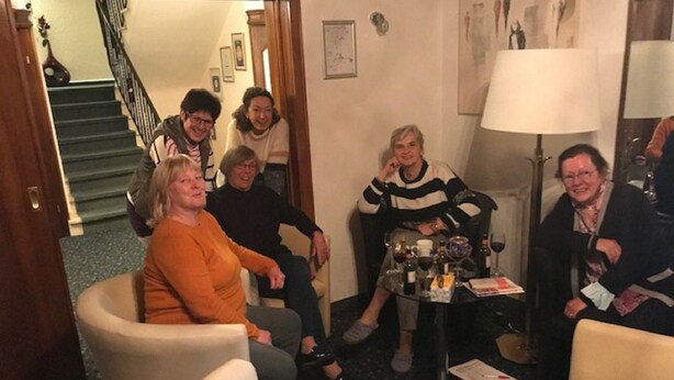 A group of people in a room with a staircase in the background, a table with bottles and glasses, and a lamp. - Hotel Schick (Oberursel)