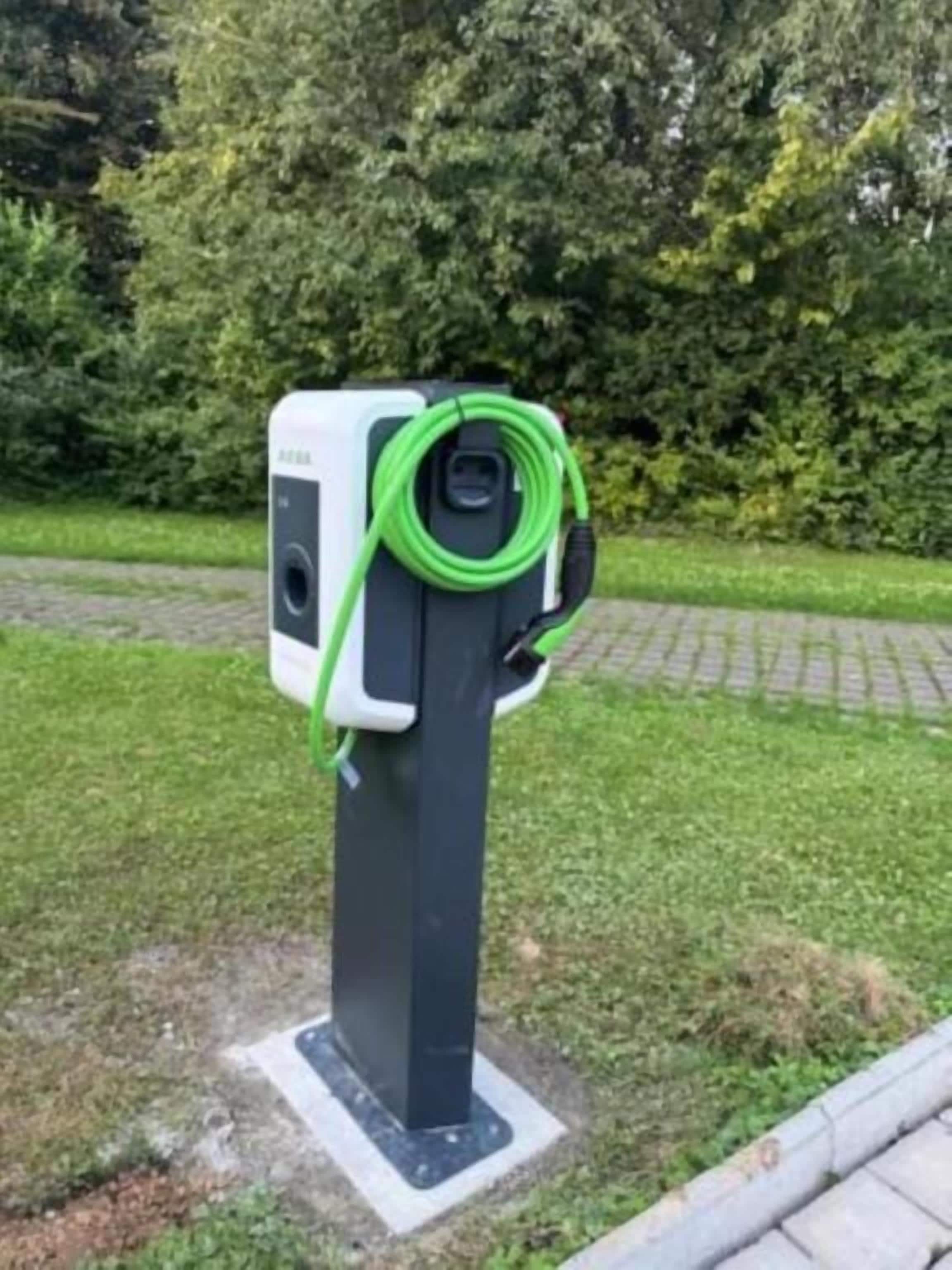Una estación de carga eléctrica fija con un cable conectado, ubicada en una zona pavimentada con césped y árboles de fondo.