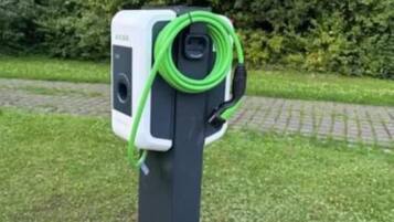 Una estación de carga eléctrica fija con un cable conectado, ubicada en una zona pavimentada con césped y árboles de fondo.