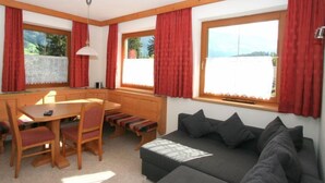 A cozy room with a wooden bench seating area, a sofa, and a dining table with chairs. Large windows with red curtains provide views of the outdoors. - Herbert by Interhome (Kaltenbach)
