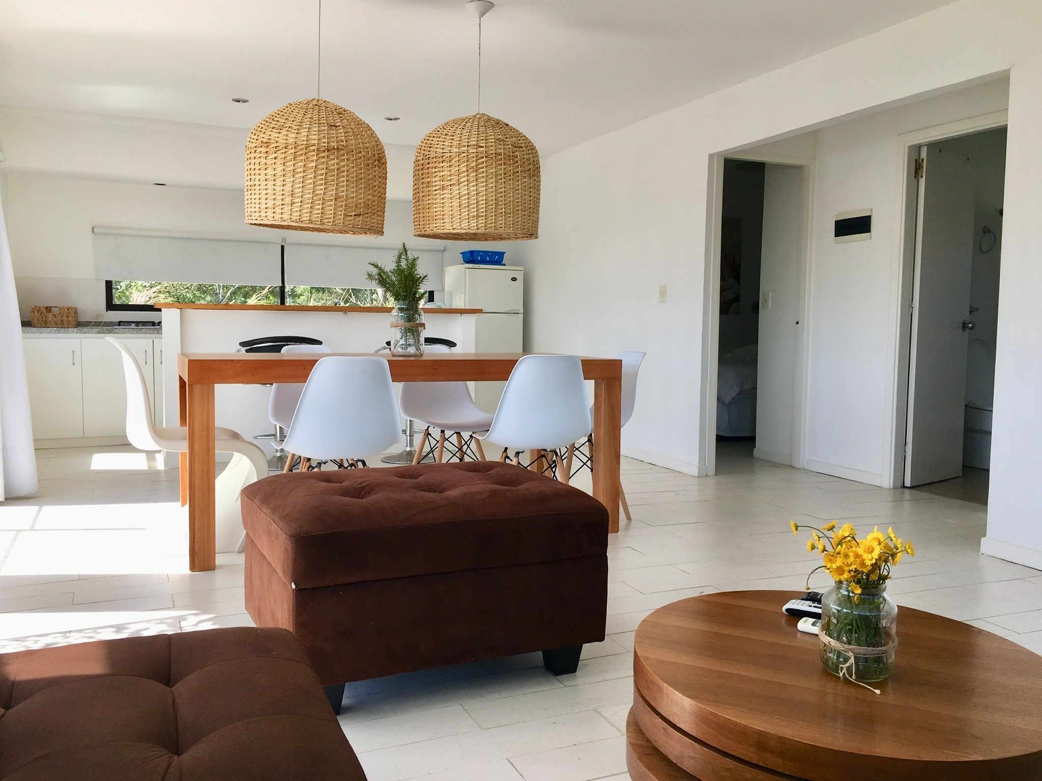 Una sala de estar moderna con un sofá marrón, una mesa de centro de madera, y un comedor con sillas blancas y una mesa de madera.