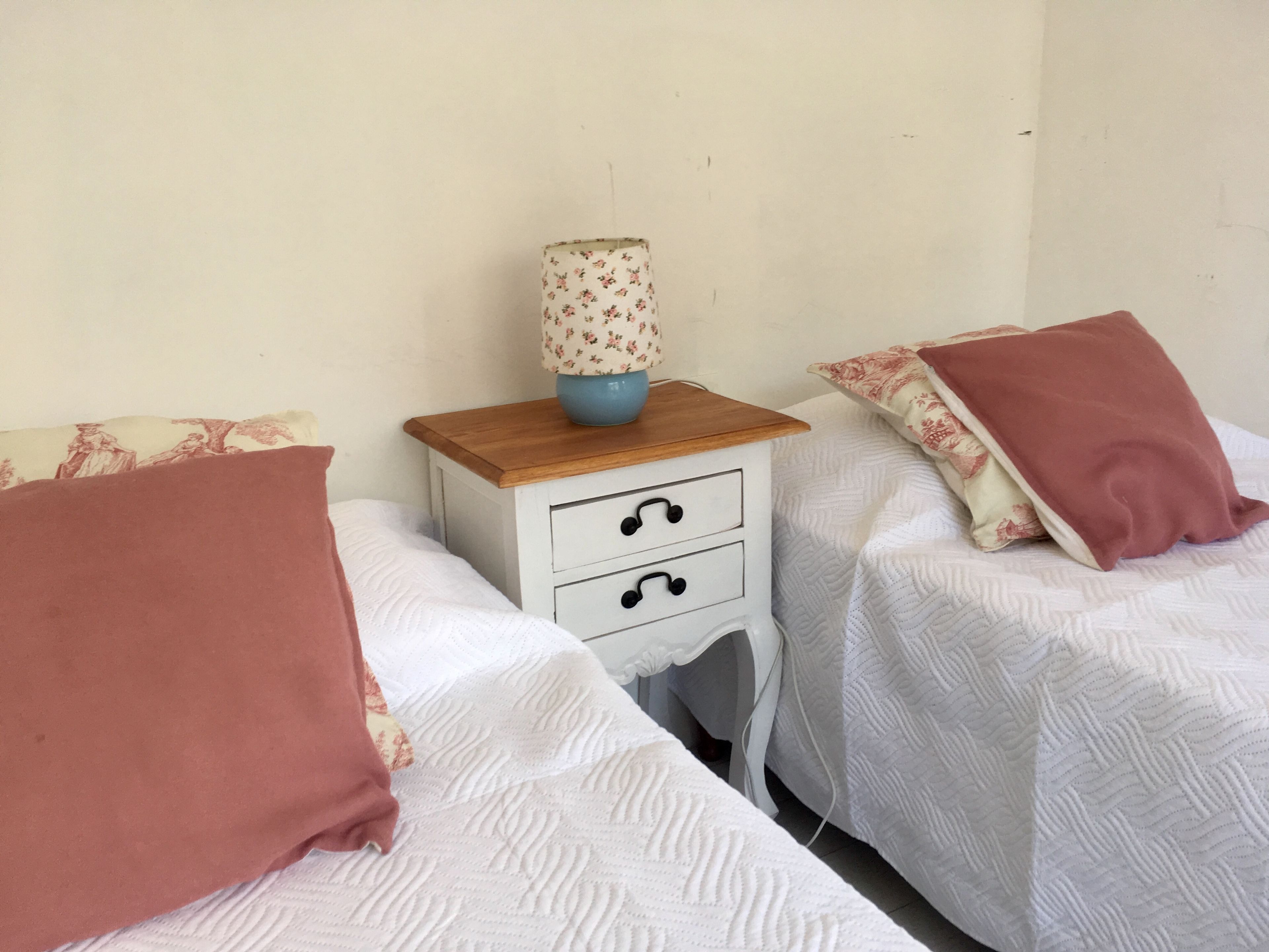 Un dormitorio con dos camas individuales, cada una con cubrecama blanco y almohada rosa. Hay una mesita de noche con una lámpara entre las camas.