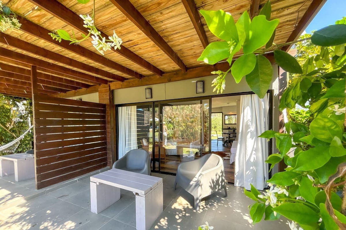 Un espace extérieur aménagé en terrasse, avec un plafond en bois, une table blanche, deux chaises et une porte vitrée donnant sur un espace intérieur.