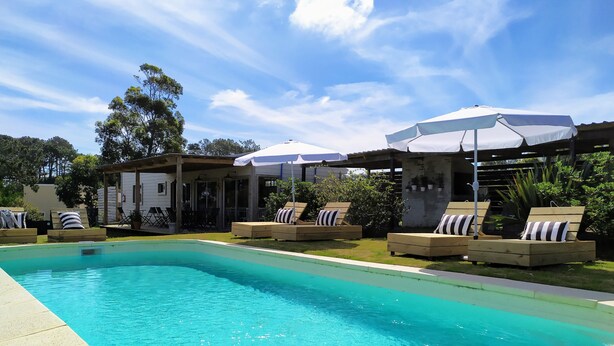 Une piscine entourée de verdure, avec des chaises longues et des parasols, sous un ciel dégagé.