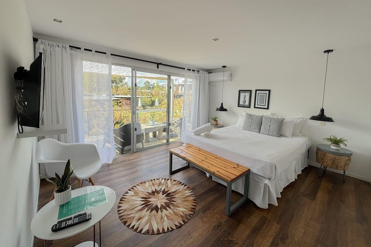 Une chambre à coucher moderne avec un grand lit, un banc en bois, une petite table avec une plante, et une vue sur l’extérieur grâce à une porte-fenêtre coulissante.