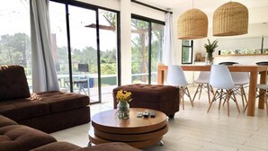 Un salon moderne avec un ensemble de canapés marron, une table basse ronde en bois ornée d’un vase de fleurs et un coin repas avec des chaises blanches et une table en bois.