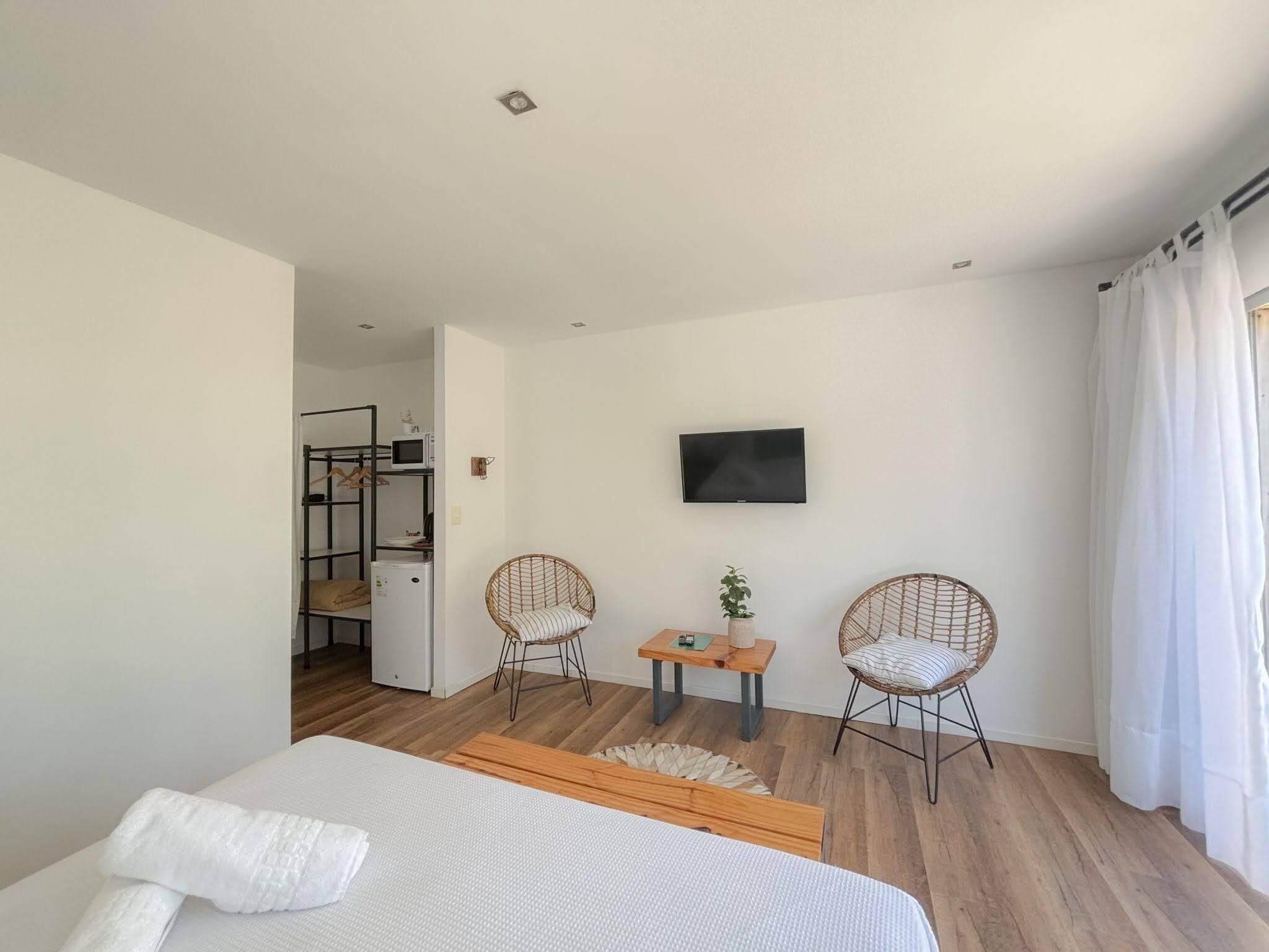 A bedroom with a bed, two wicker chairs, a small table with a plant, a TV on the wall, and a kitchenette in the background.