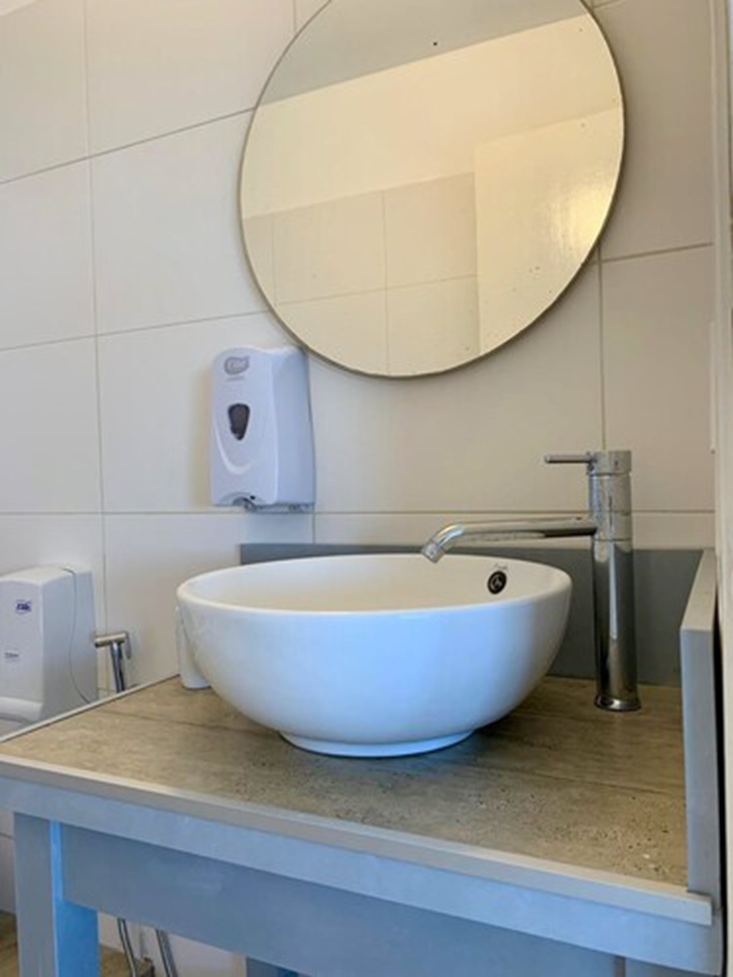 A bathroom with a round mirror, a white sink, and a soap dispenser on the wall.
