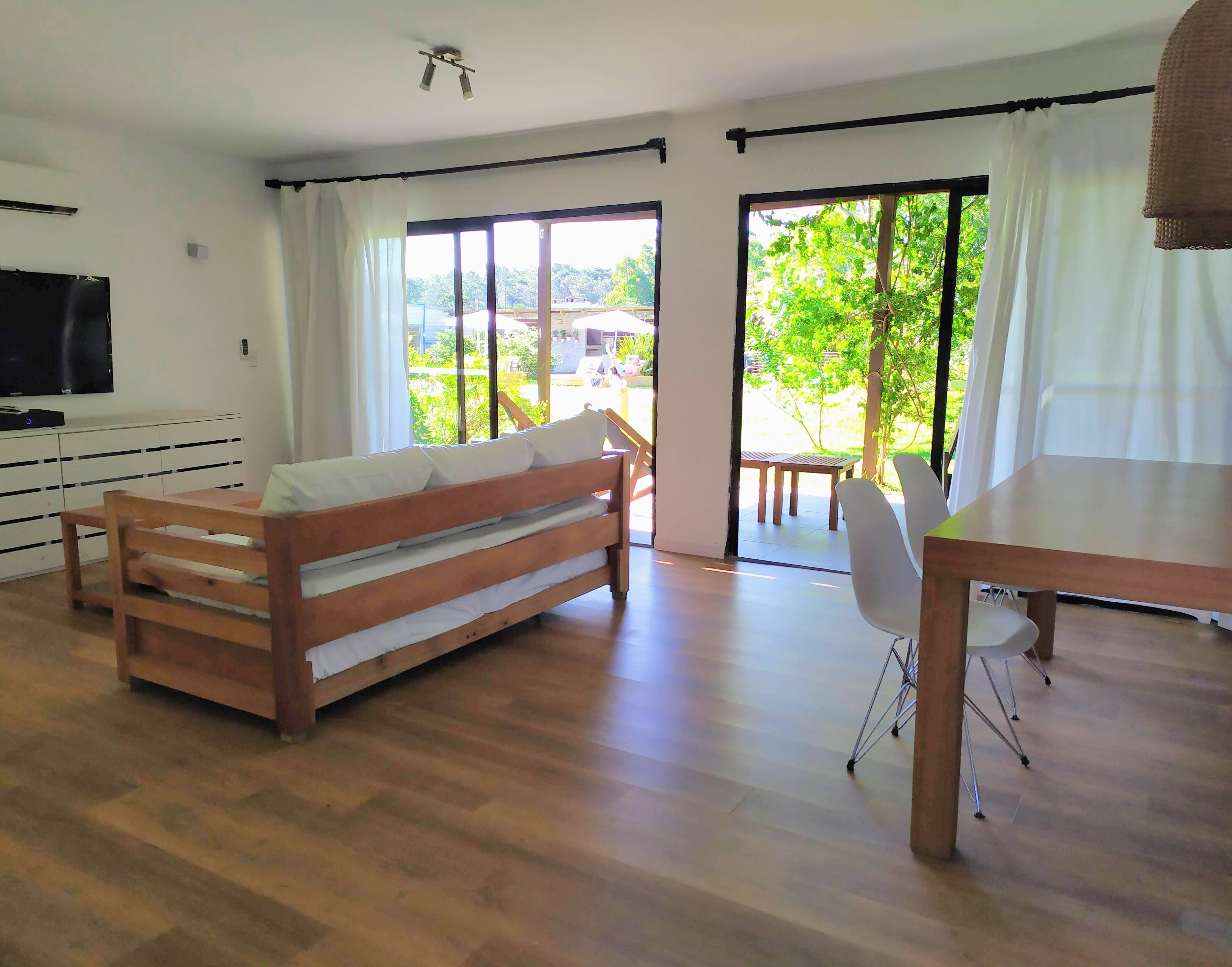 Amplia sala de estar con sofá de madera, una mesa de comedor y un televisor. Amplias ventanas ofrecen vistas al exterior.