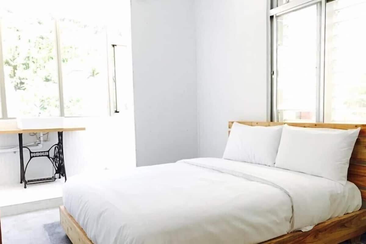 Un dormitorio con estructura de cama de madera, ropa de cama blanca y una máquina de coser en la esquina.