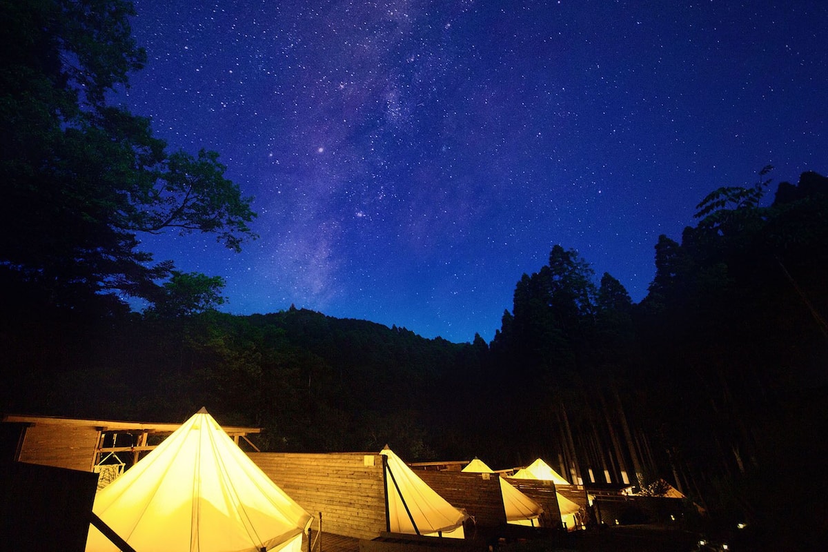 A starry night sky with a visible Milky Way, surrounded by forested hills and illuminated tents.