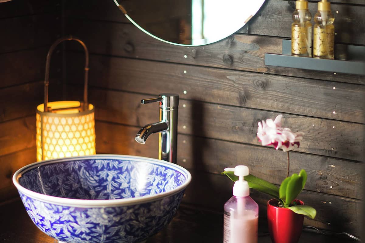 A bathroom with a blue and white patterned bowl sink, a modern faucet, a hanging towel, a lit candle, a mirror, a shelf with bottles, and a potted plant.