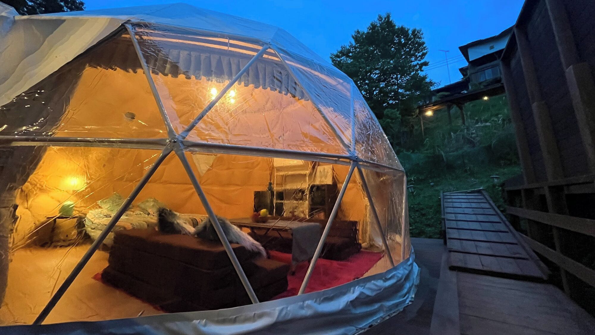 Tienda en forma de cúpula geodésica con terraza de madera, iluminación interior y vistas al bosque circundante.