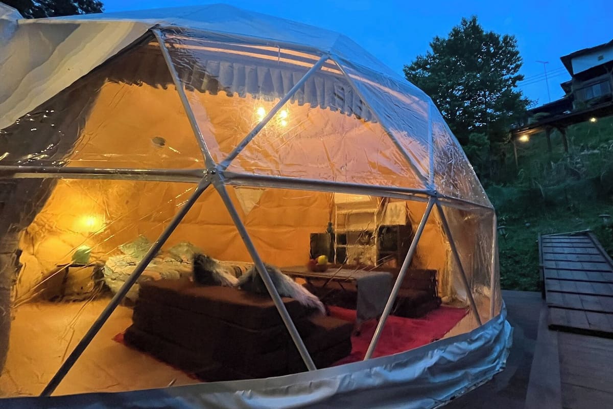 A geodesic dome tent with a wooden deck, interior lighting, and a view of the surrounding forest.