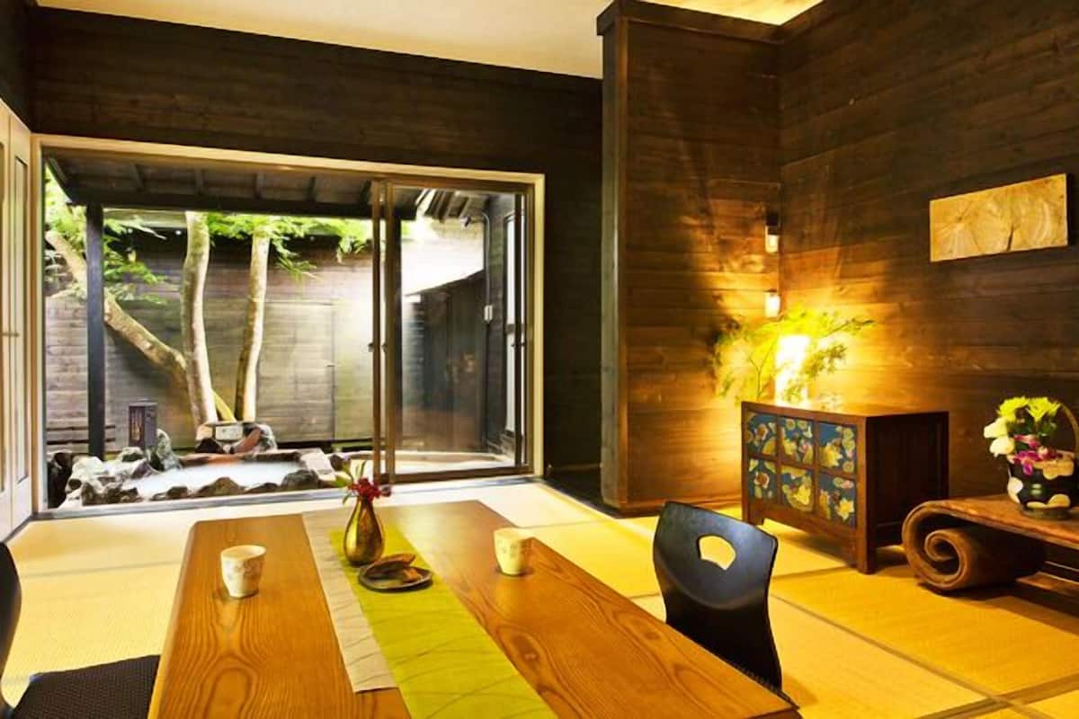 A dining area with a wooden table, chairs, a vase with flowers, and a view through a glass door to an outdoor space with trees.