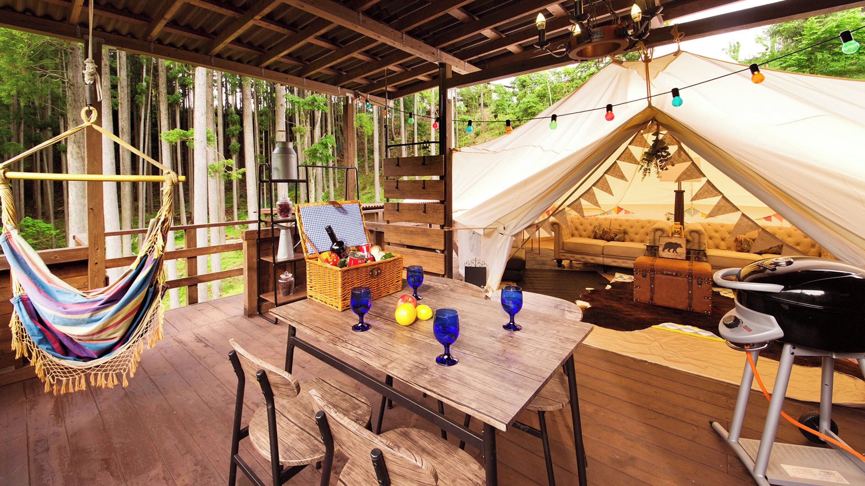 A wooden deck with a hammock, a table with blue wine glasses and a basket, a tent with a sofa inside, and a grill outside.