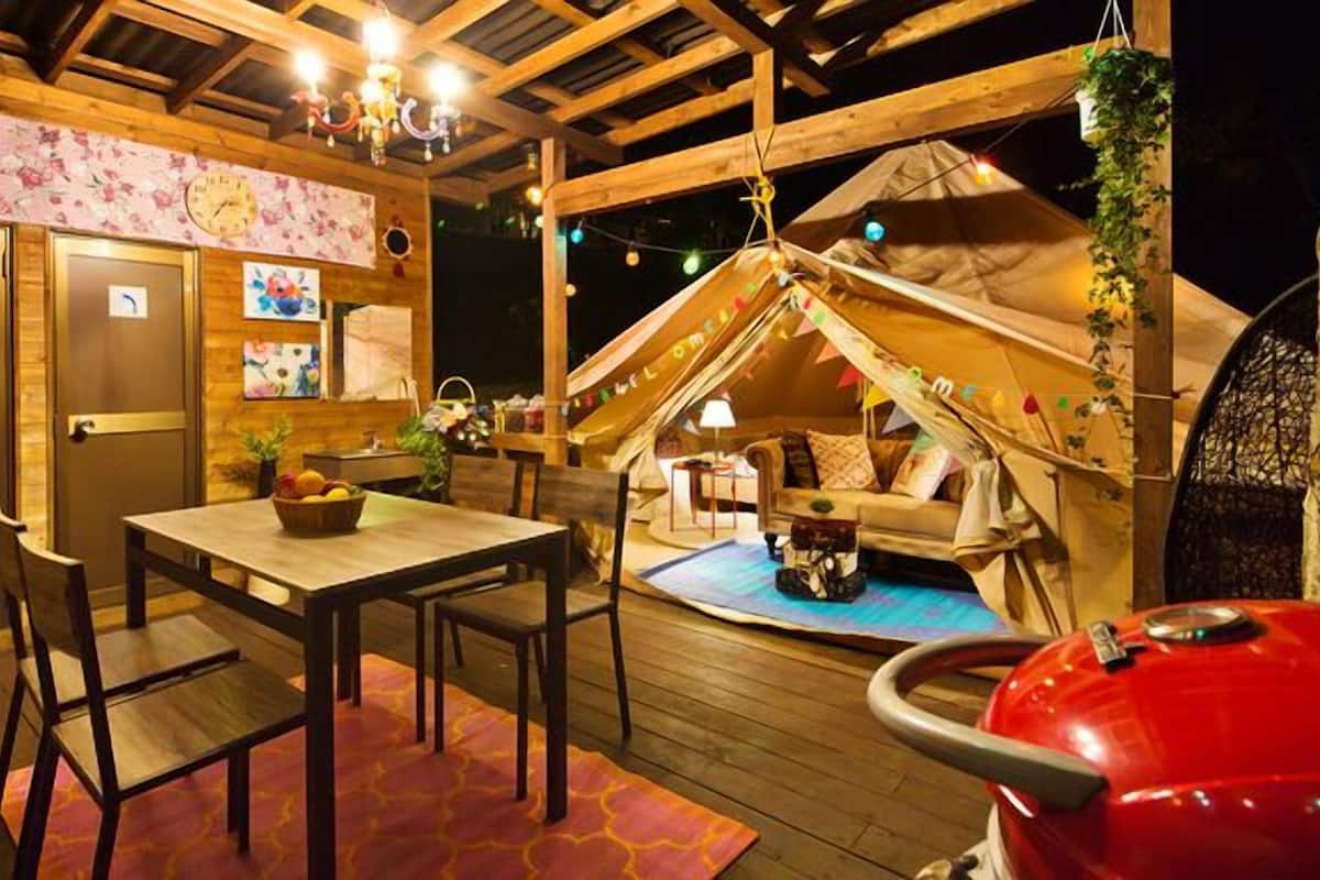 A cozy outdoor dining area with a wooden table and chairs, a red grill, and a tent-like structure with a 'Welcome' sign.