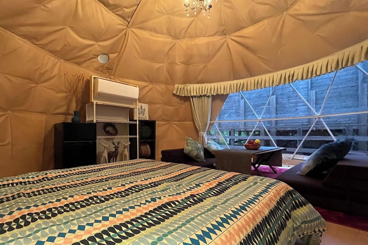 A tented room with a bed, a small table with a bowl of fruit, and a window with a view of a building.