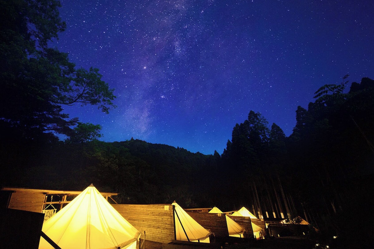 A starry night sky with a visible Milky Way, surrounded by forested hills and illuminated tents.
