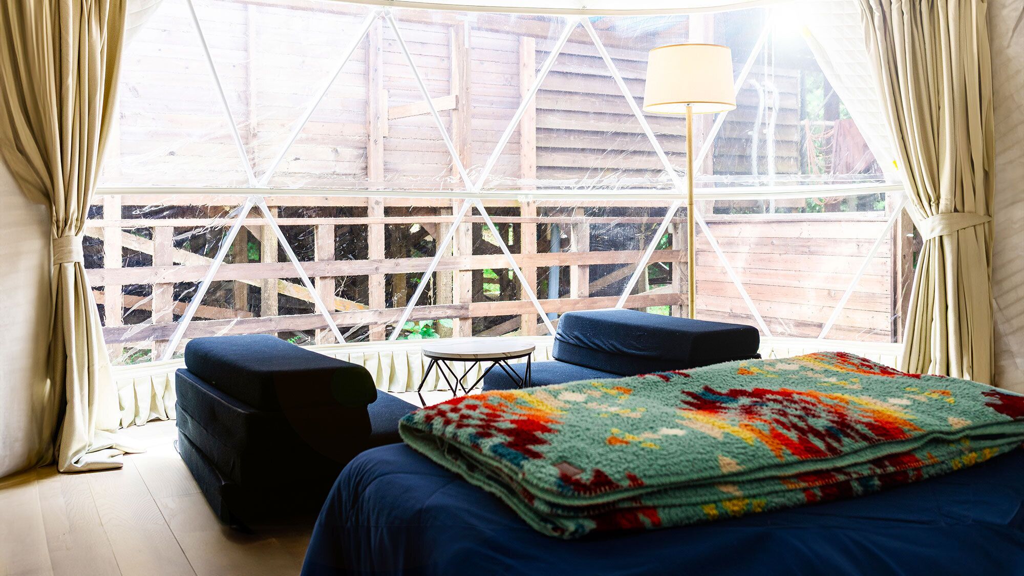 Una habitació amb un llit, una taula petita, dues cadires, una làmpada i vista a través d'una finestra de l'atrezzo de fusta.