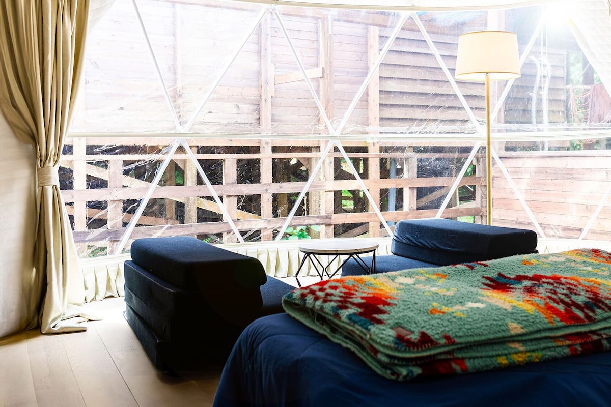 A room with a bed, a small table, two chairs, a lamp, and a view through a wooden lattice window.