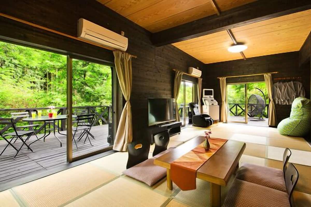 A wooden interior with tatami mats, a television, a massage chair, and a large window overlooking greenery.