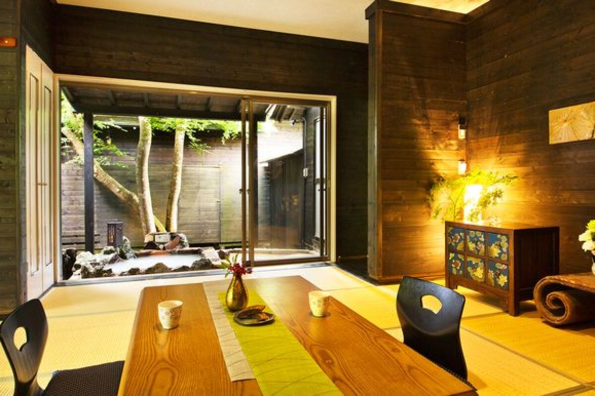 A dining area with a wooden table, chairs, a vase with flowers, and a view through a glass door to an outdoor space with trees.