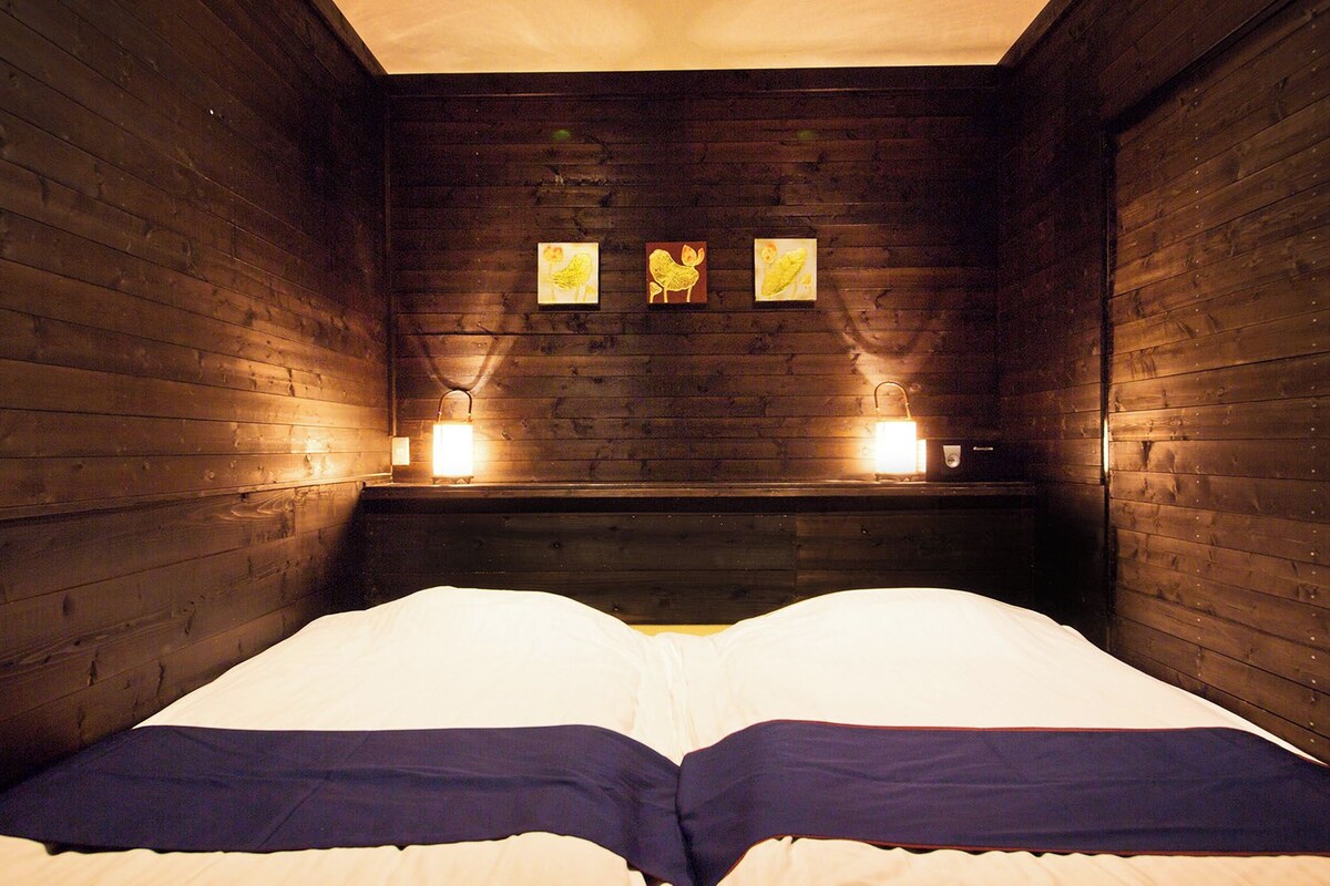 A wooden room with a bed, two wall-mounted lights, and three framed pictures.