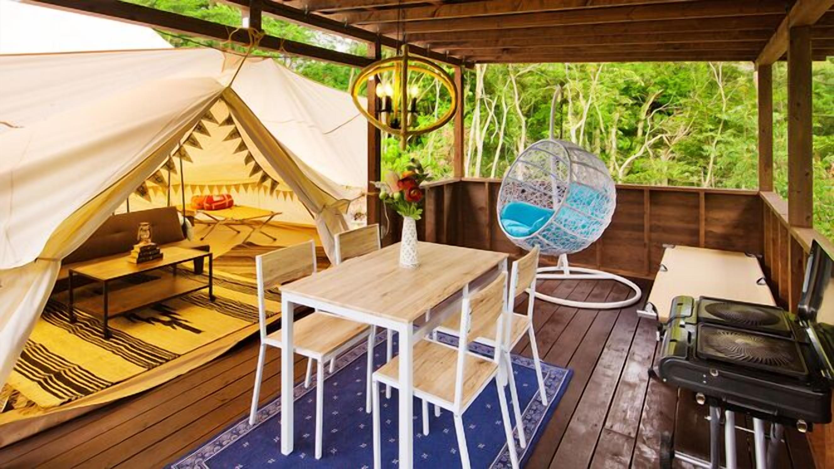 Alojamiento con carpa, mesa de comedor, sillas, una silla colgante y un ventilador.