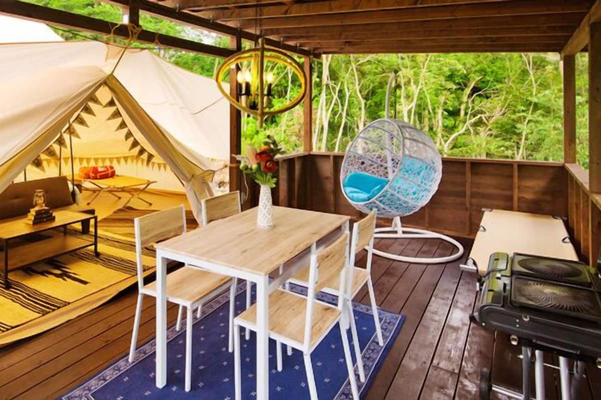 A tented accommodation with a dining table, chairs, a hanging chair, and a fan.