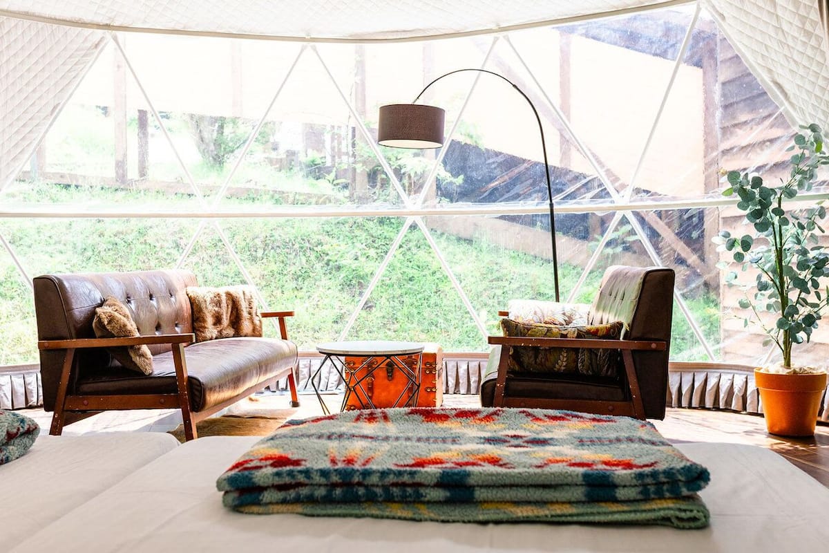 A cozy room with a bed, two chairs, a small table, and a large window with a view of greenery.