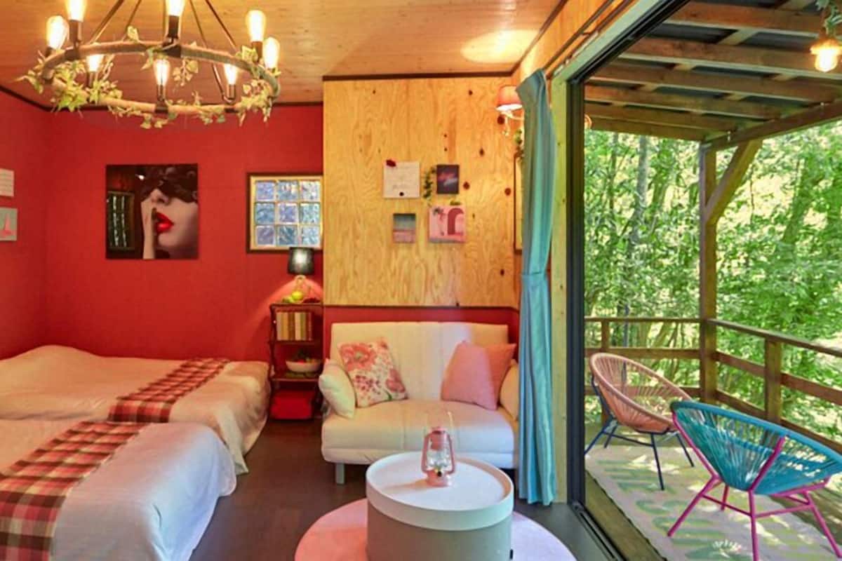 A room with two beds, a round table, a chair, and a balcony with a view of greenery.